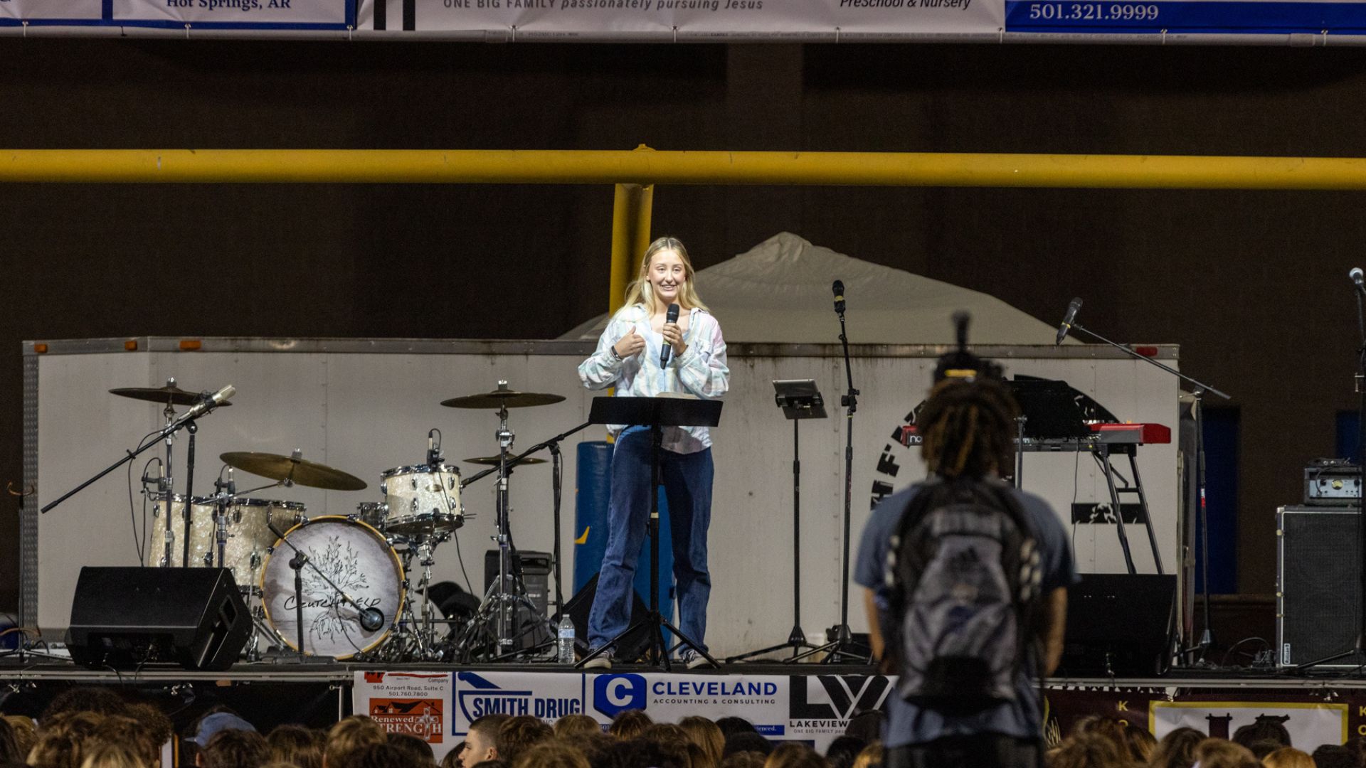 A young woman with blonde hair stands at a podium on a stage, speaking into a microphone. She is dressed in a light-colored top and jeans, and is smiling as she addresses an audience