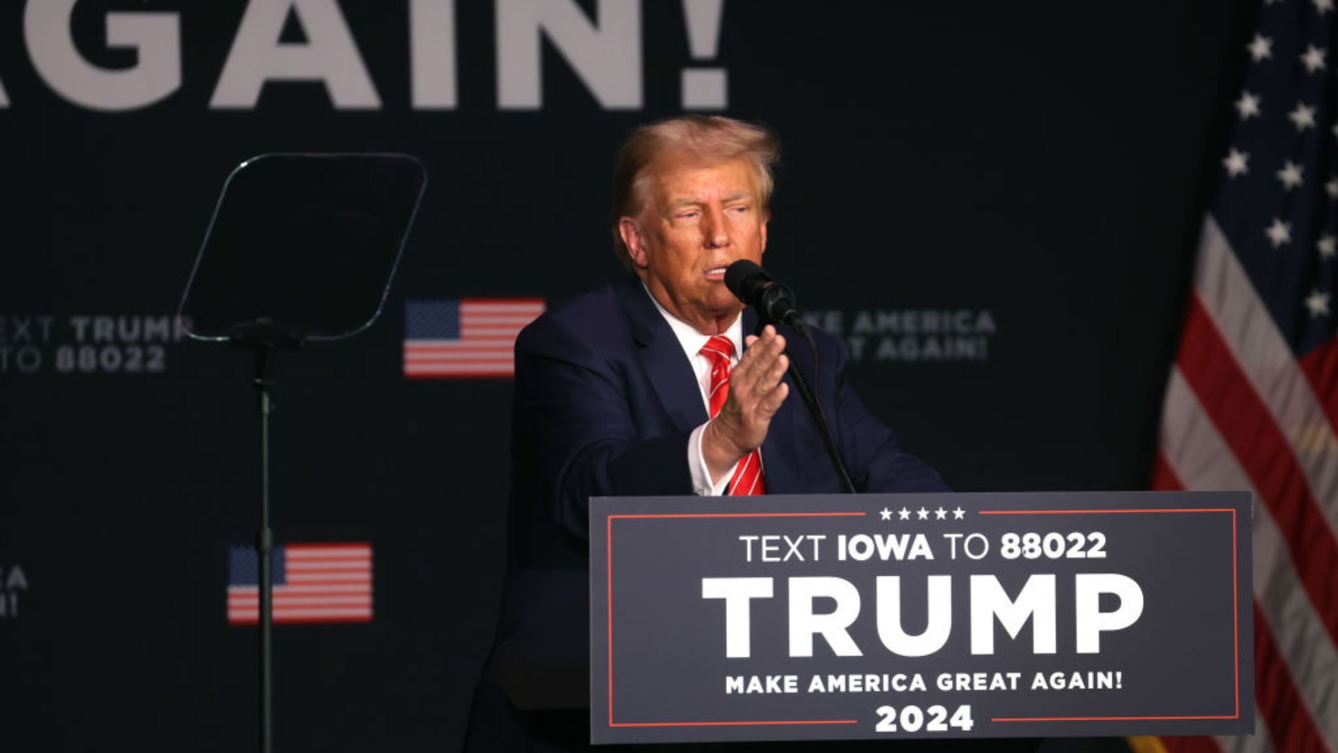 Former President Donald Trump speaking at a podium during a campaign-style event, with promotional banners in the background