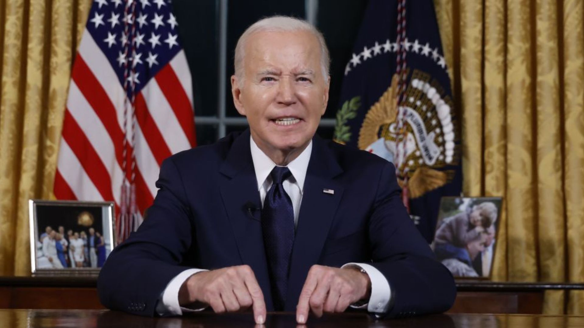 President Joe Biden, wearing a navy suit, sits at the Oval Office desk with the American flag and the presidential seal flag in the background