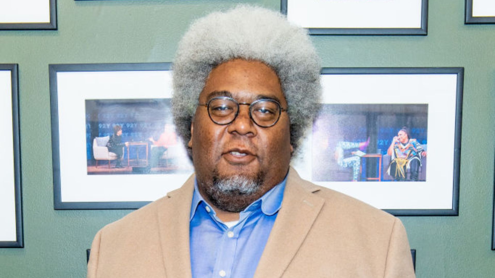 Elie Mystal wearing a beige blazer and blue shirt, standing in front of a green wall adorned with framed photos