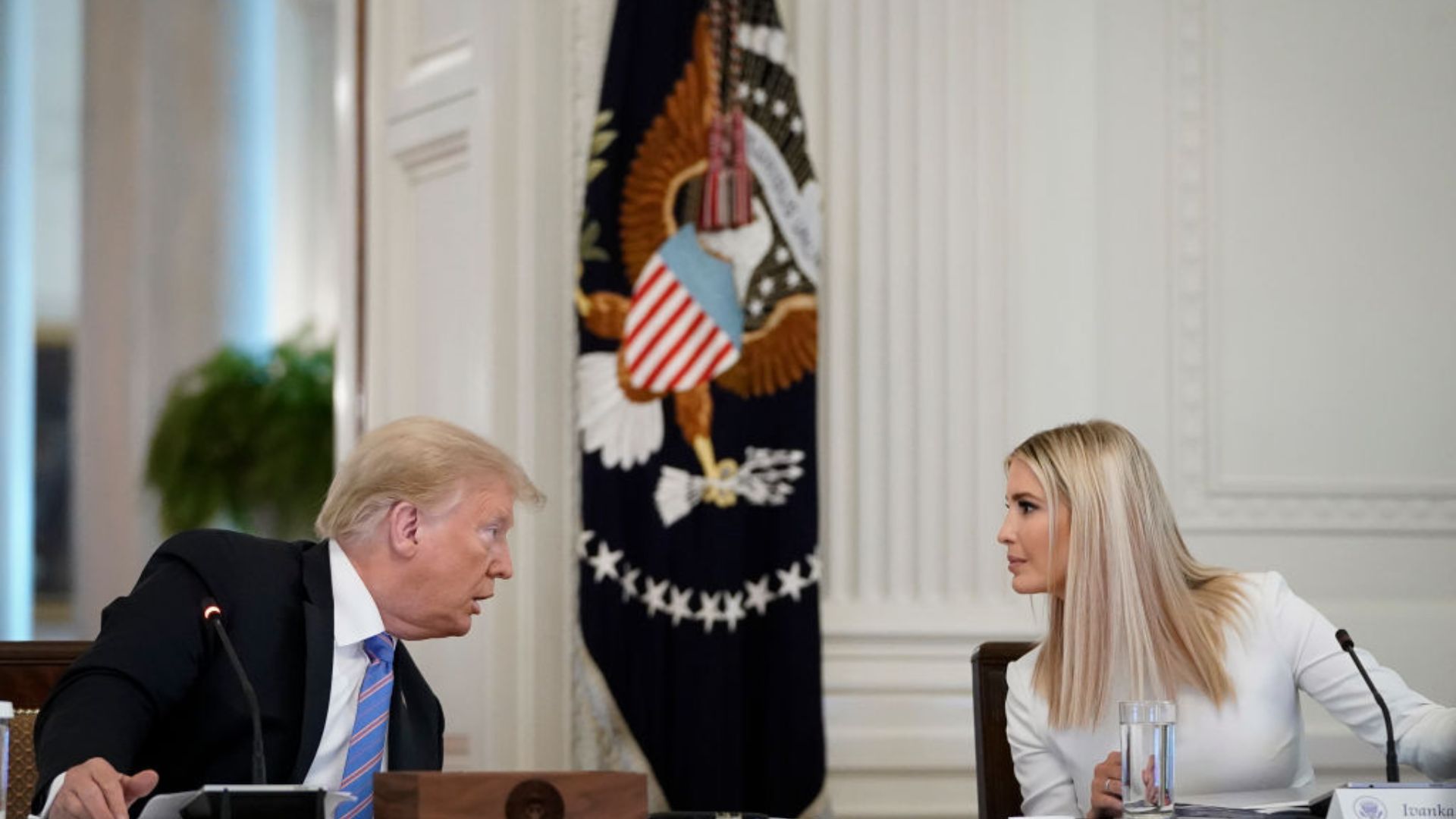 Donald Trump, wearing a dark suit and blue tie, sits to the left of the image and turns his head to his left with a focused expression. To the right, Ivanka Trump, wearing a white blouse, looks to her left while seated at a table with a glass of water in front of her