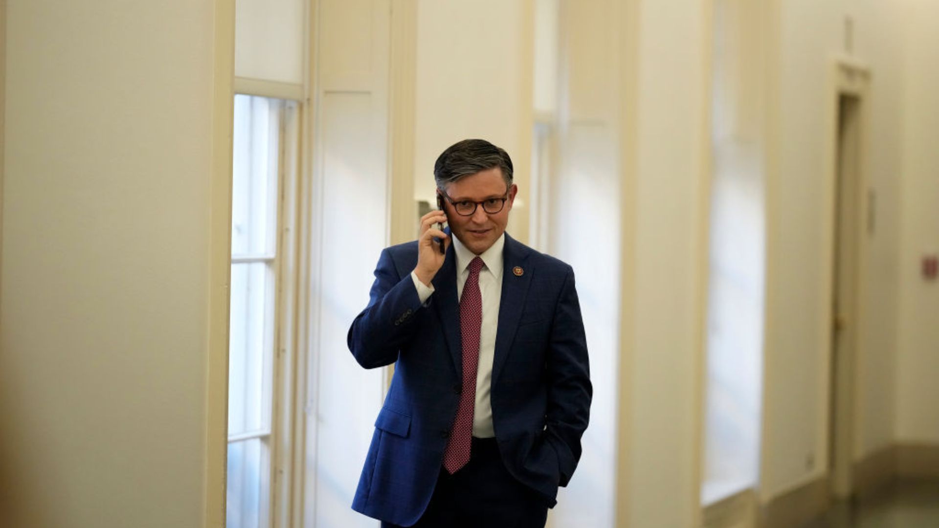 Mike Johnson, wearing glasses, a blue suit jacket, and a red tie, talks on a cell phone