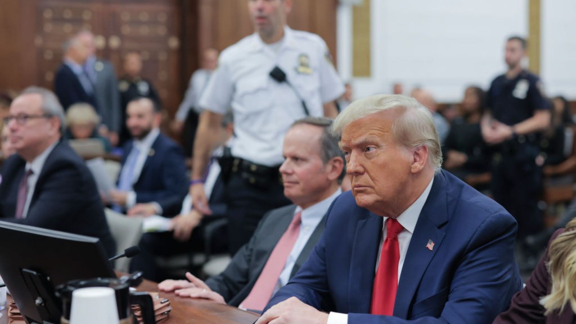 Donald Trump, wearing a navy suit and red tie, seated in a courtroom. He looks intently forward with a focused expression