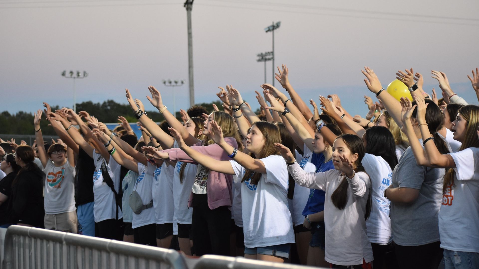 A group of diverse individuals, predominantly young, stand closely together outdoors near a metal barrier. Many of them are wearing white or light-colored shirts. They're raising their hands high