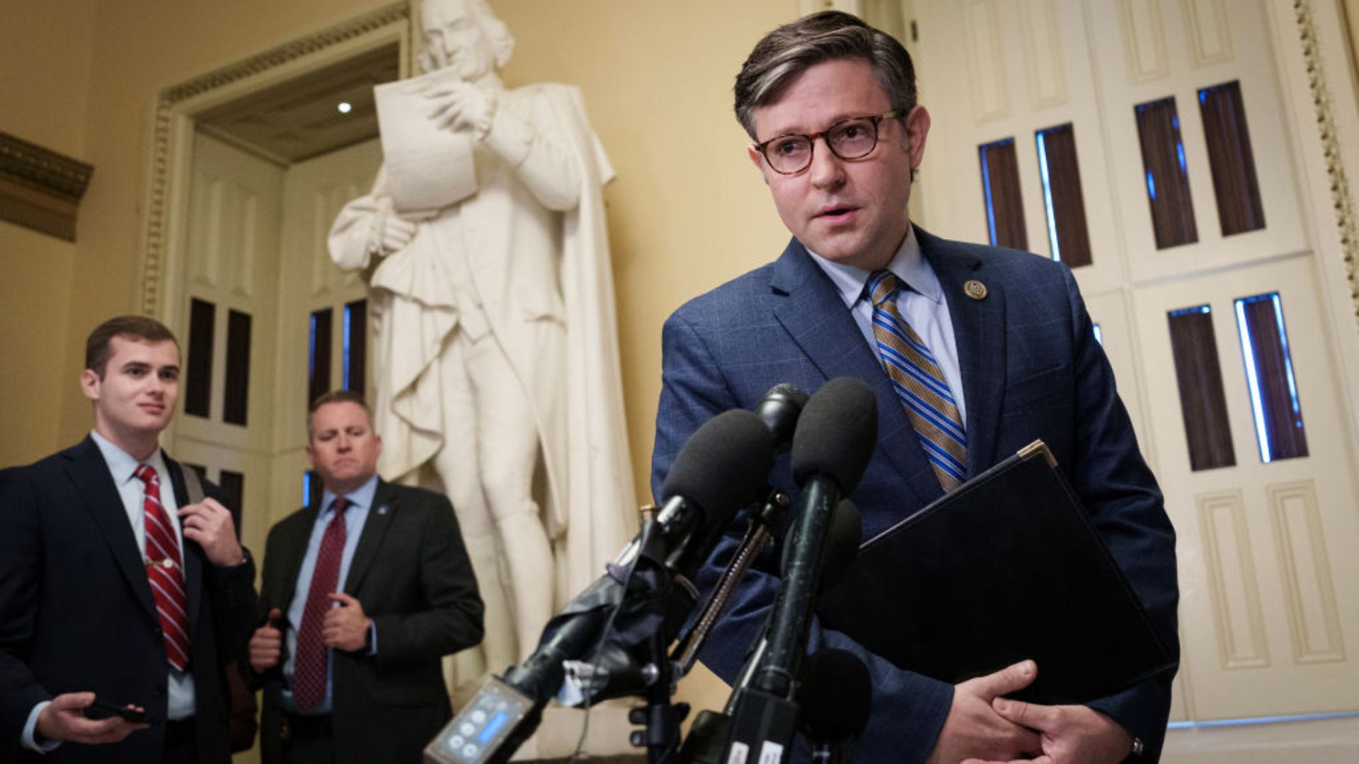 Mike Johnson, wearing a blue suit and glasses, speaks into microphones in a grand room. Behind him stands a statue of a historical figure and two men