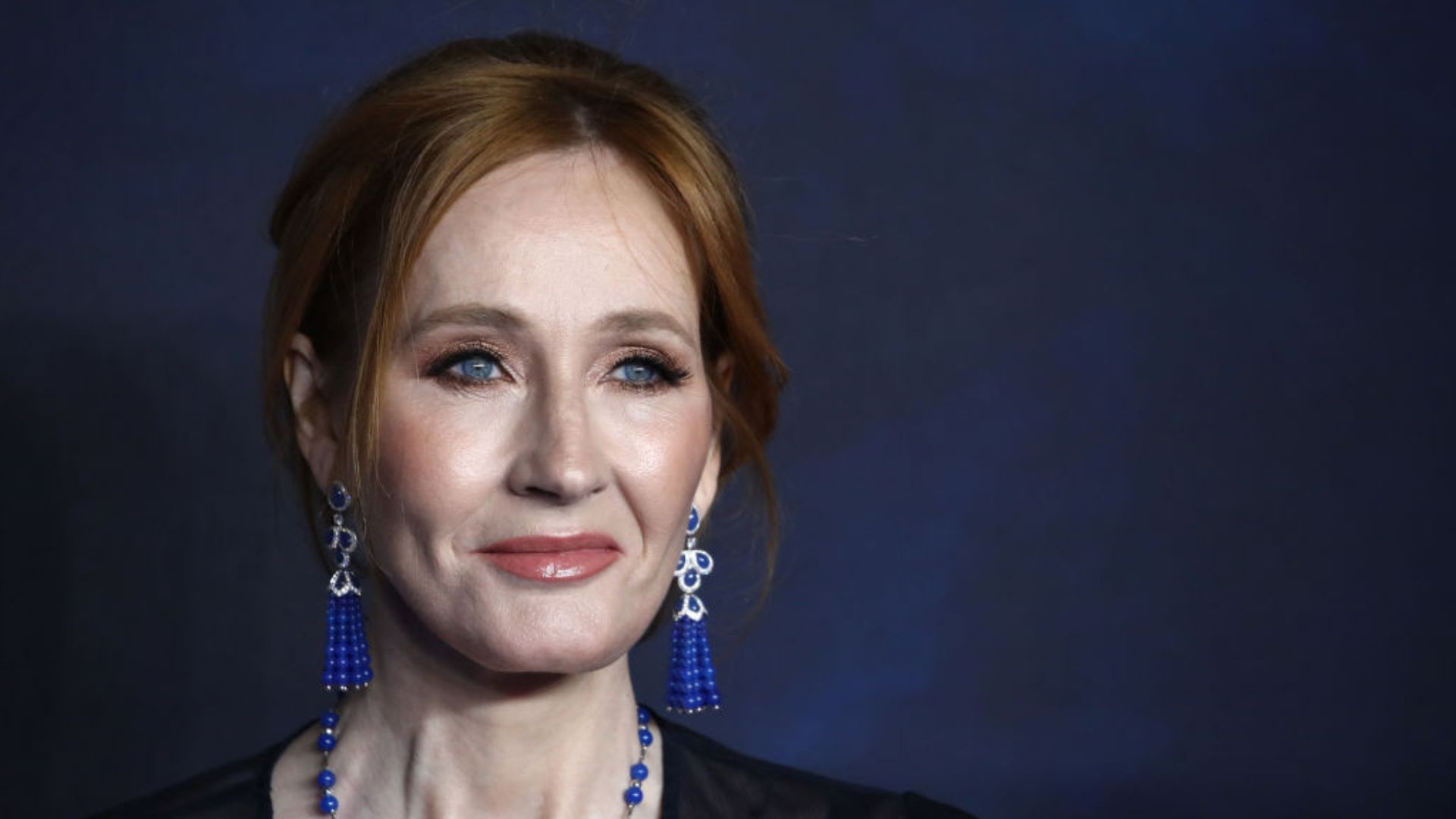 Portrait of J.K. Rowling at a public event. She is photographed from the shoulders up, wearing a black outfit and blue earrings