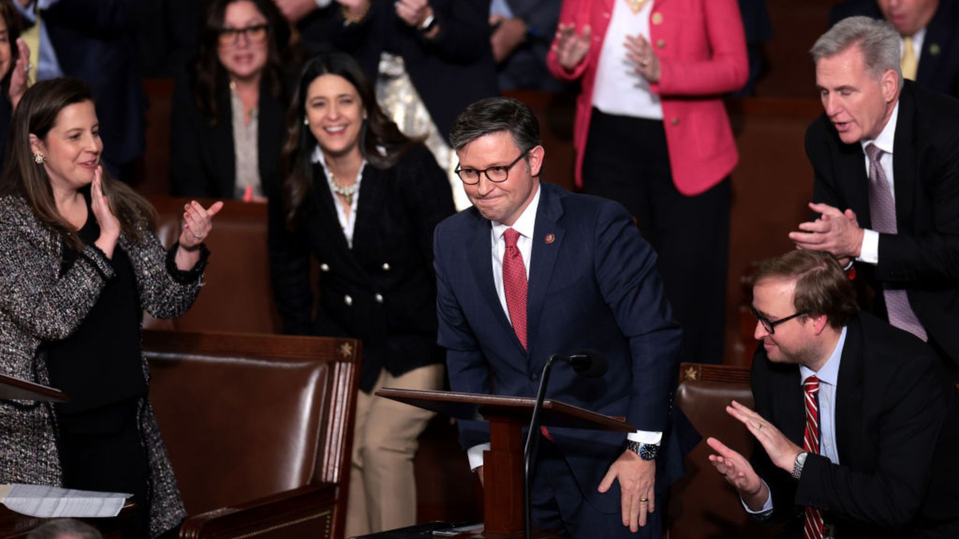 Mike Johnson, wearing glasses and a dark suit with a red tie, is standing behind a podium with his right hand on some papers, receiving applause from people around him in a formal setting