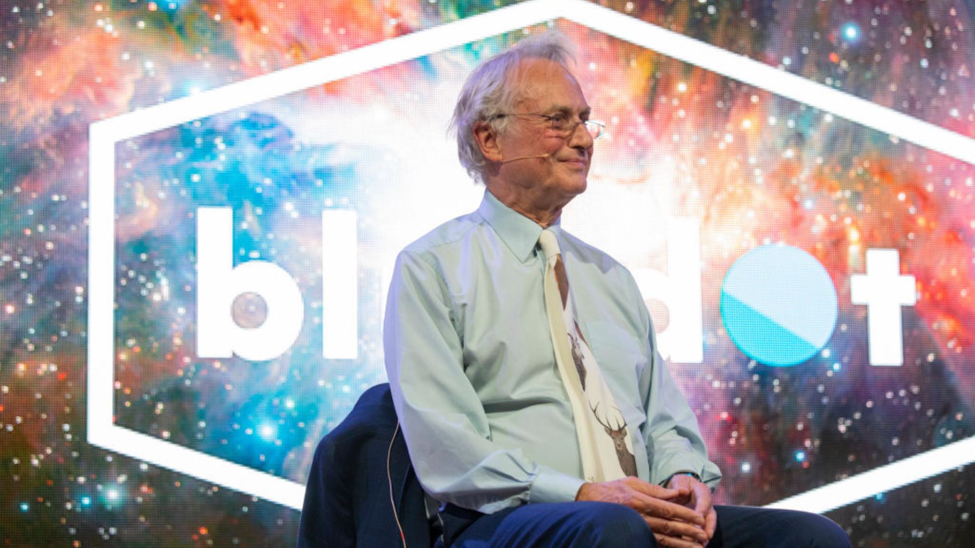 Richard Dawkins seated at an event, with a colorful cosmic background