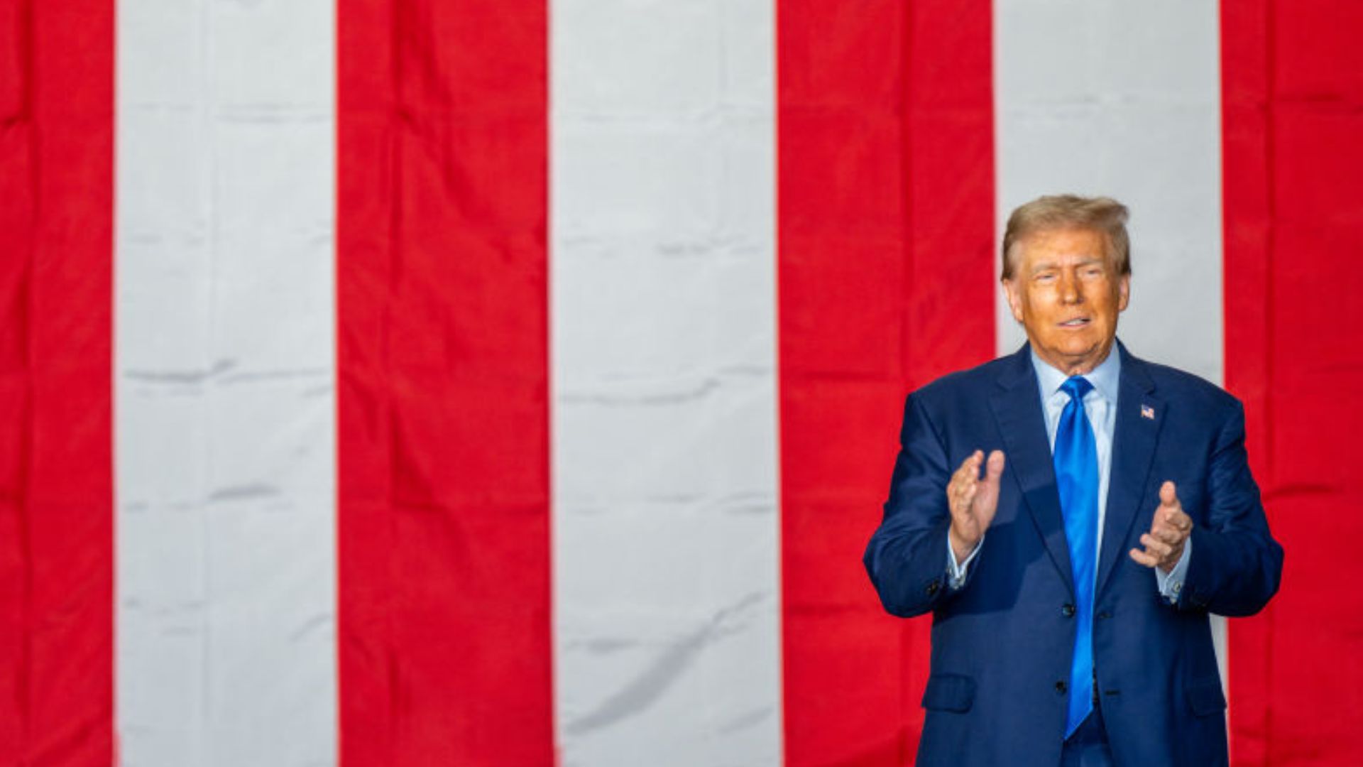 Former President Donald Trump wearing a blue suit and red tie, standing and speaking in front of a large American flag