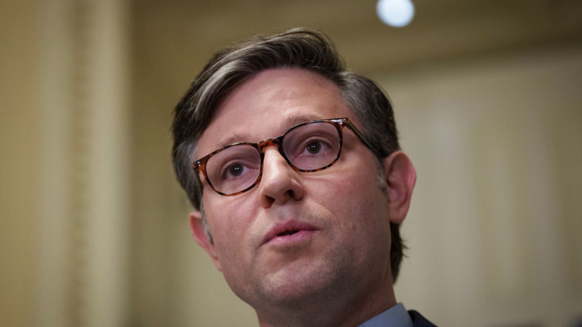 A close-up image of Mike Johnson, wearing glasses, speaking in front of a beige background