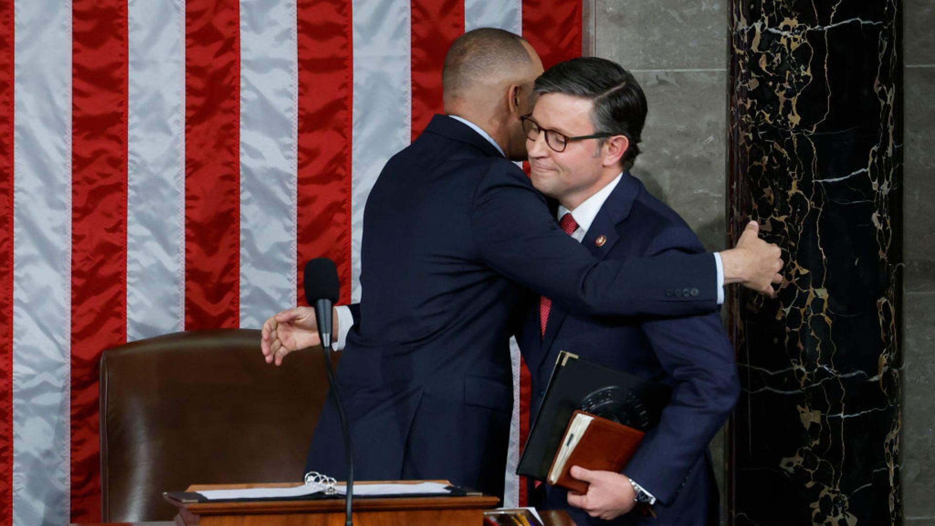 Mike Johnson, wearing glasses and a dark suit, is in a half-embrace with another person in a suit