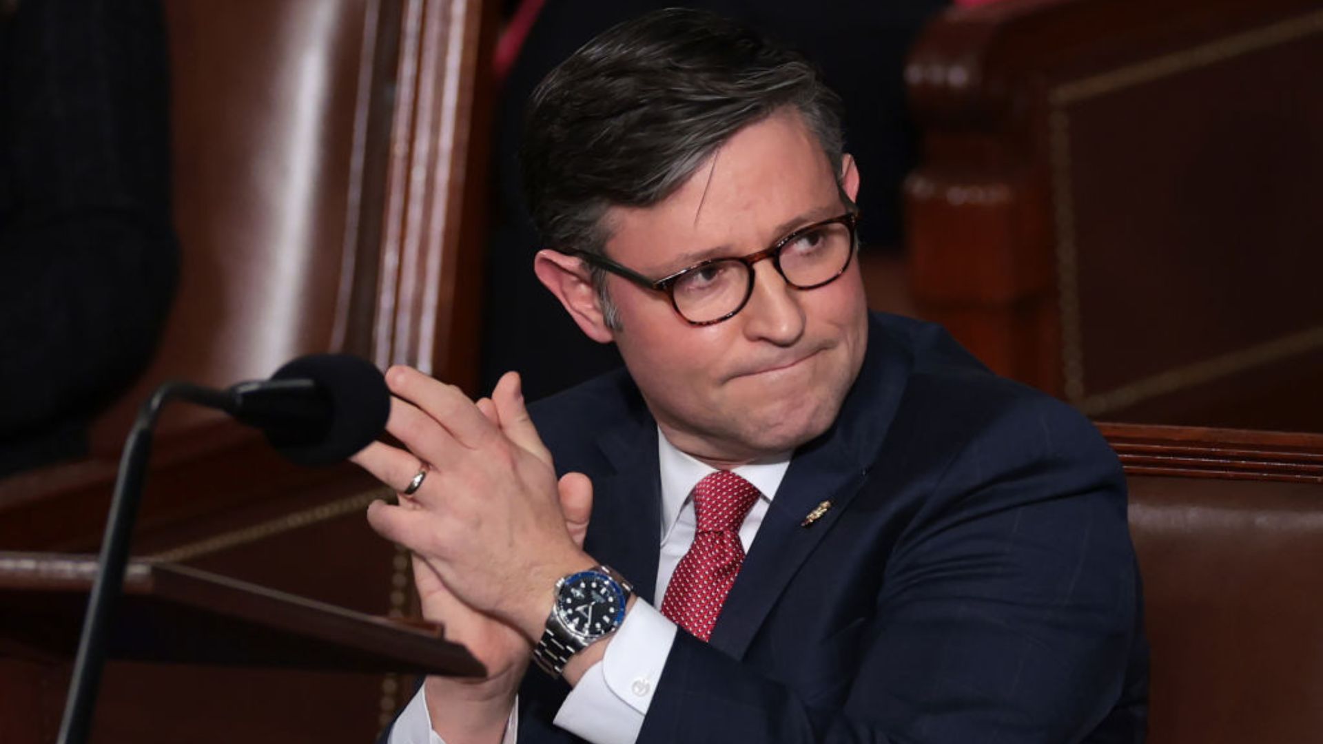Mike Johnson, wearing glasses, a dark suit, and a red tie, clapping his hands in a room with wooden interiors