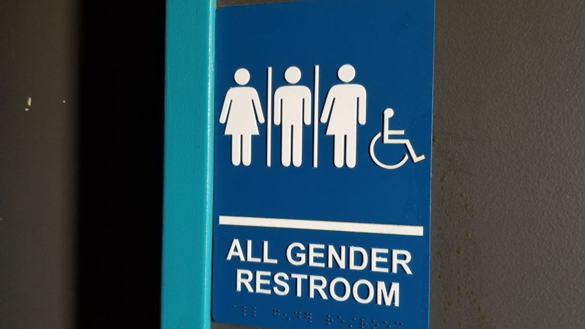 A sign on a wall indicating an 'All Gender Restroom' with pictograms of a woman, a person with no gender indication, a man, and a wheelchair symbol for accessibility