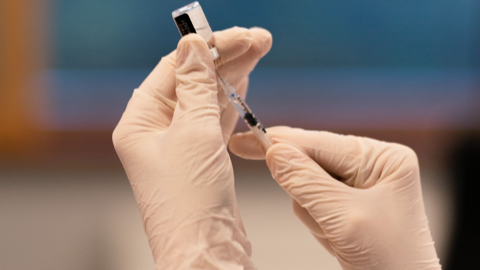Hands encased in latex gloves carefully handling a syringe and a vial, preparing a vaccine dose against a softly focused background
