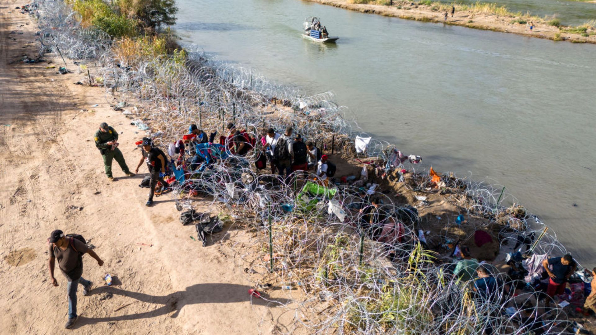 An aerial view of a group of people gathered near a body of water with razor wire barriers. A uniformed person is seen interacting with someone