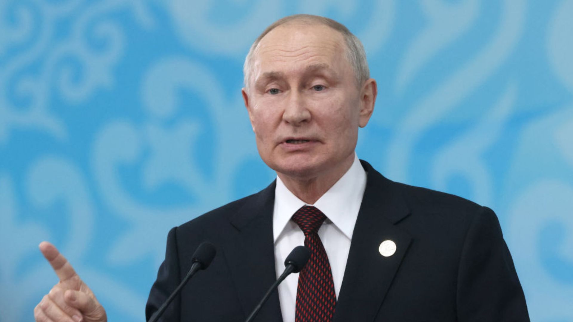 Vladimir Putin, the President of Russia, dressed in a suit and tie, speaking and gesturing with his right hand.